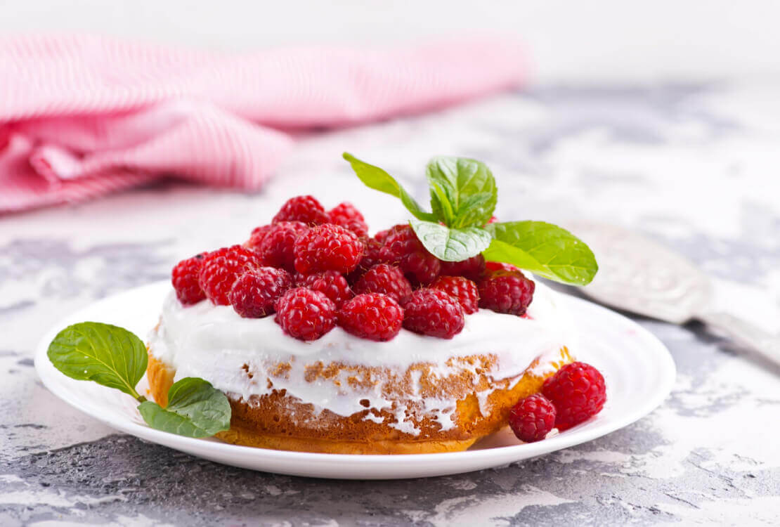 Minitaartje met wit glazuur, verse frambozen en muntblaadjes op een wit bord.