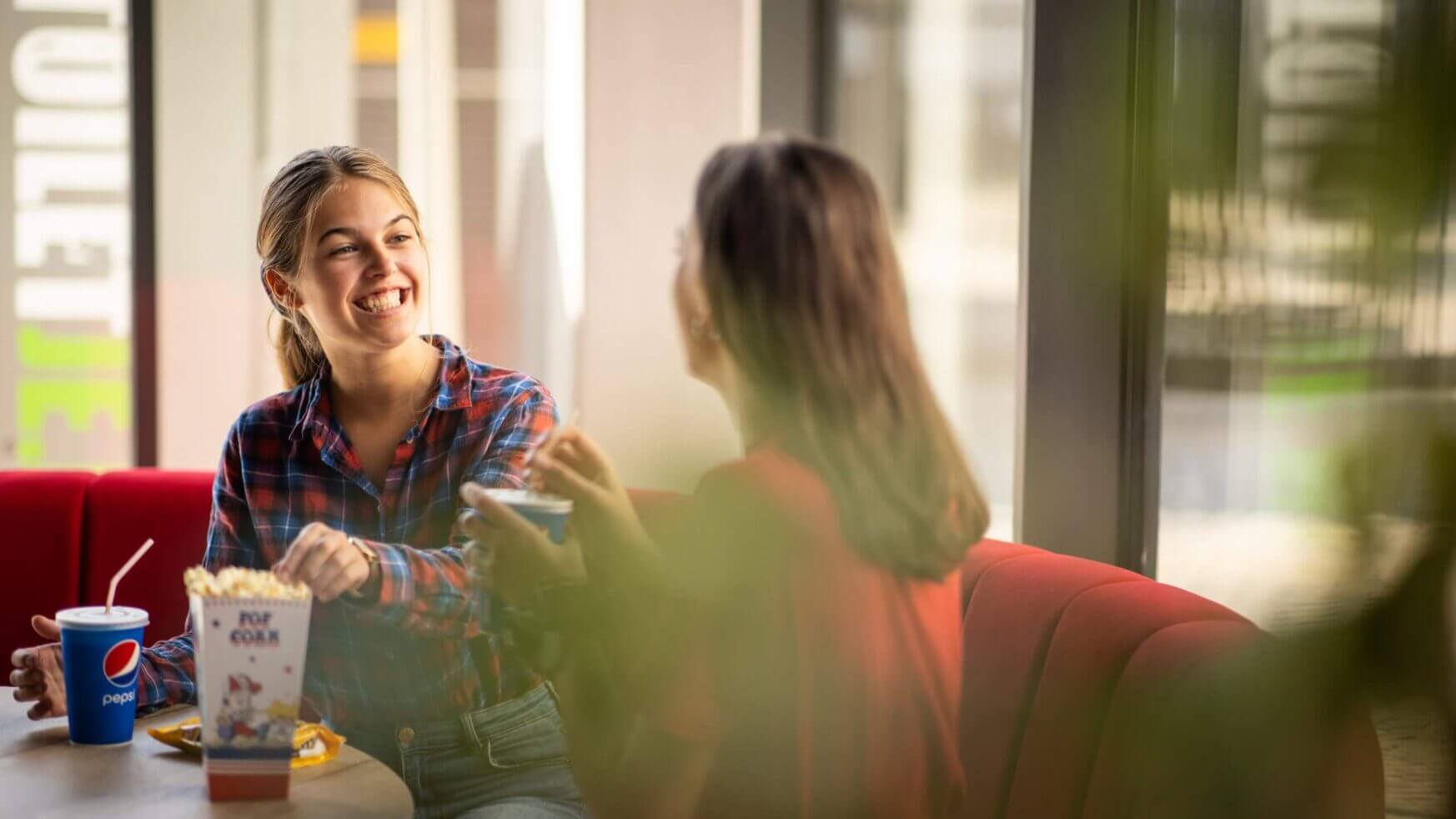 Twee vrouwen lachen en kletsen gezellig aan een zonnig cafétafeltje, onder het genot van popcorn en een drankje.