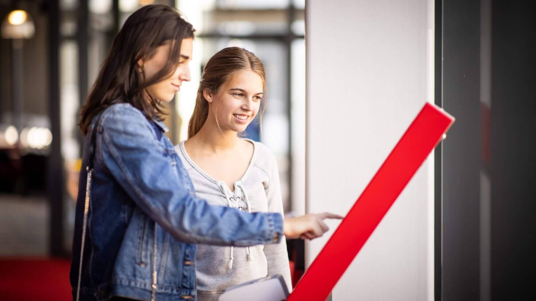 Twee vrouwen werken samen bij een touchscreen-kiosk in een lichte binnenruimte.