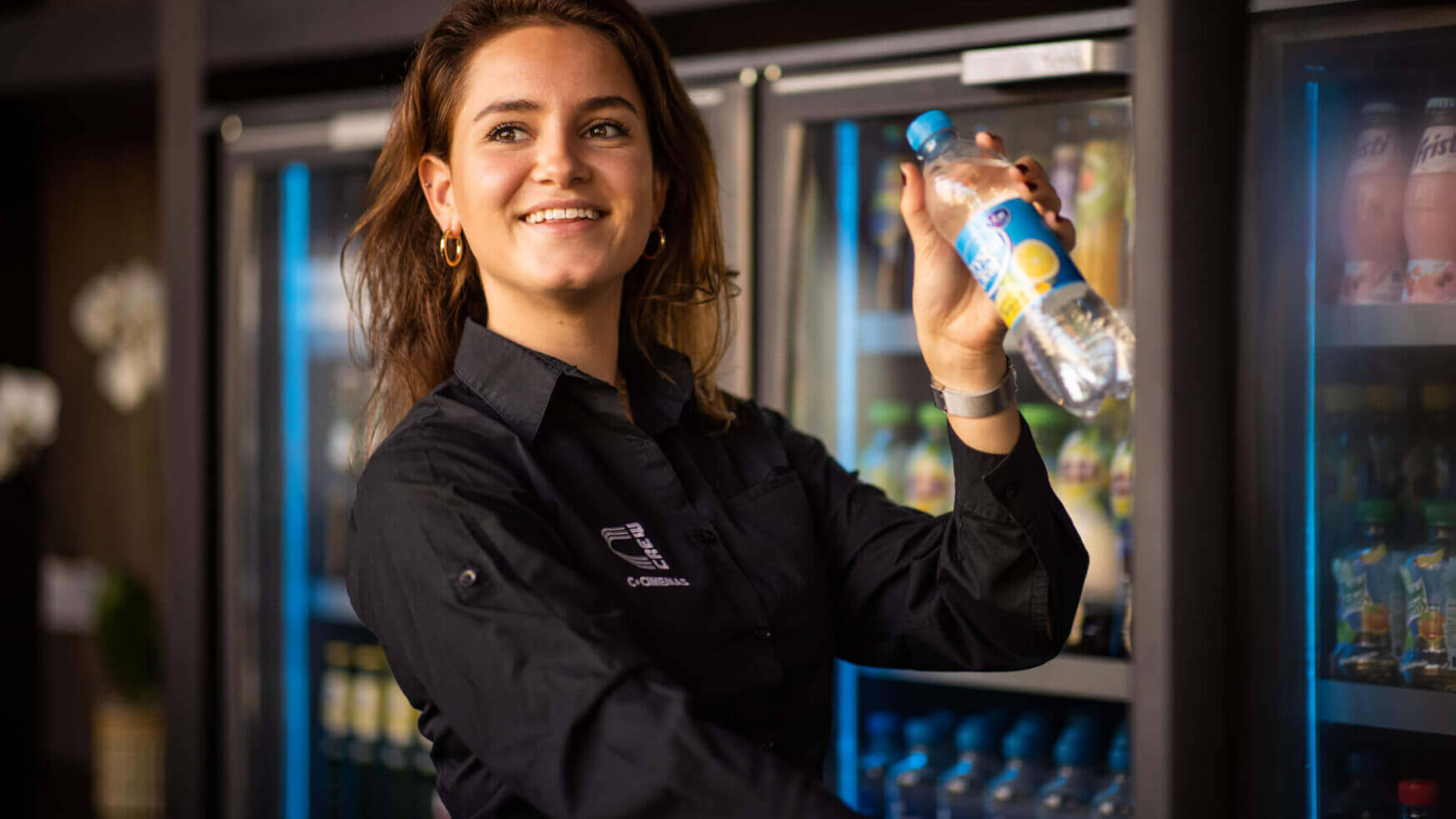 Vrouw in zwart shirt lacht met fles in haar hand bij de koelkast.