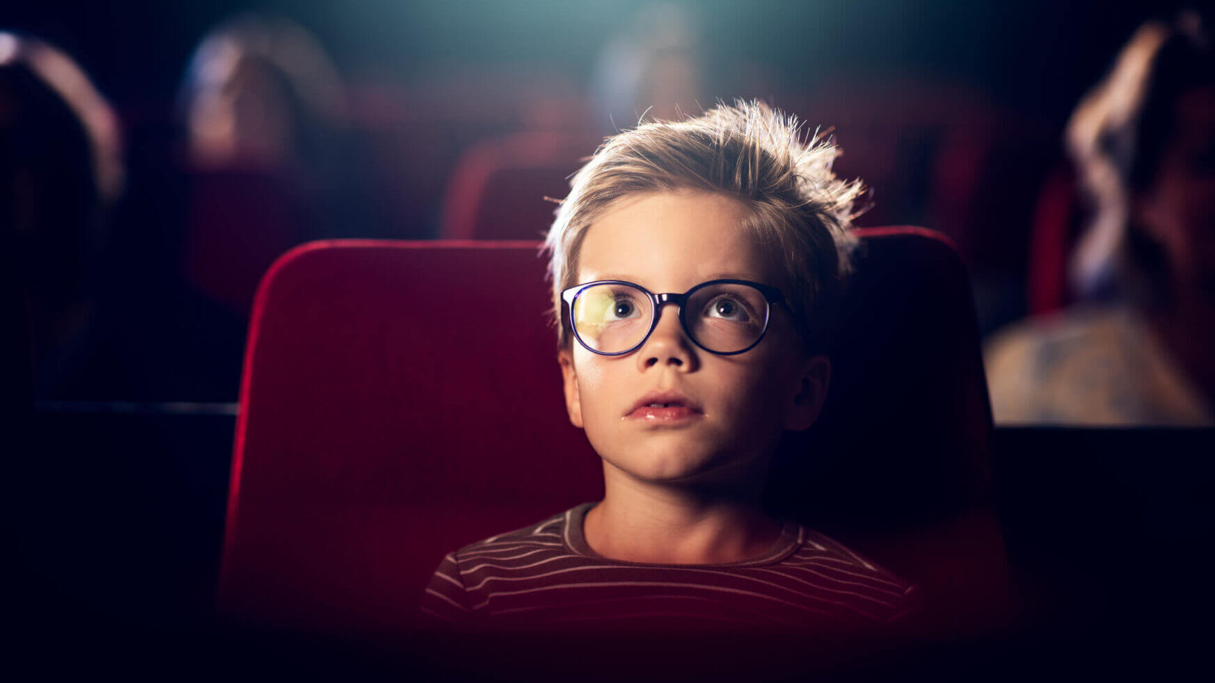 Een jongen met een bril kijkt in een schemerige bioscoopzaal naar een film, zittend op rode bekleding.