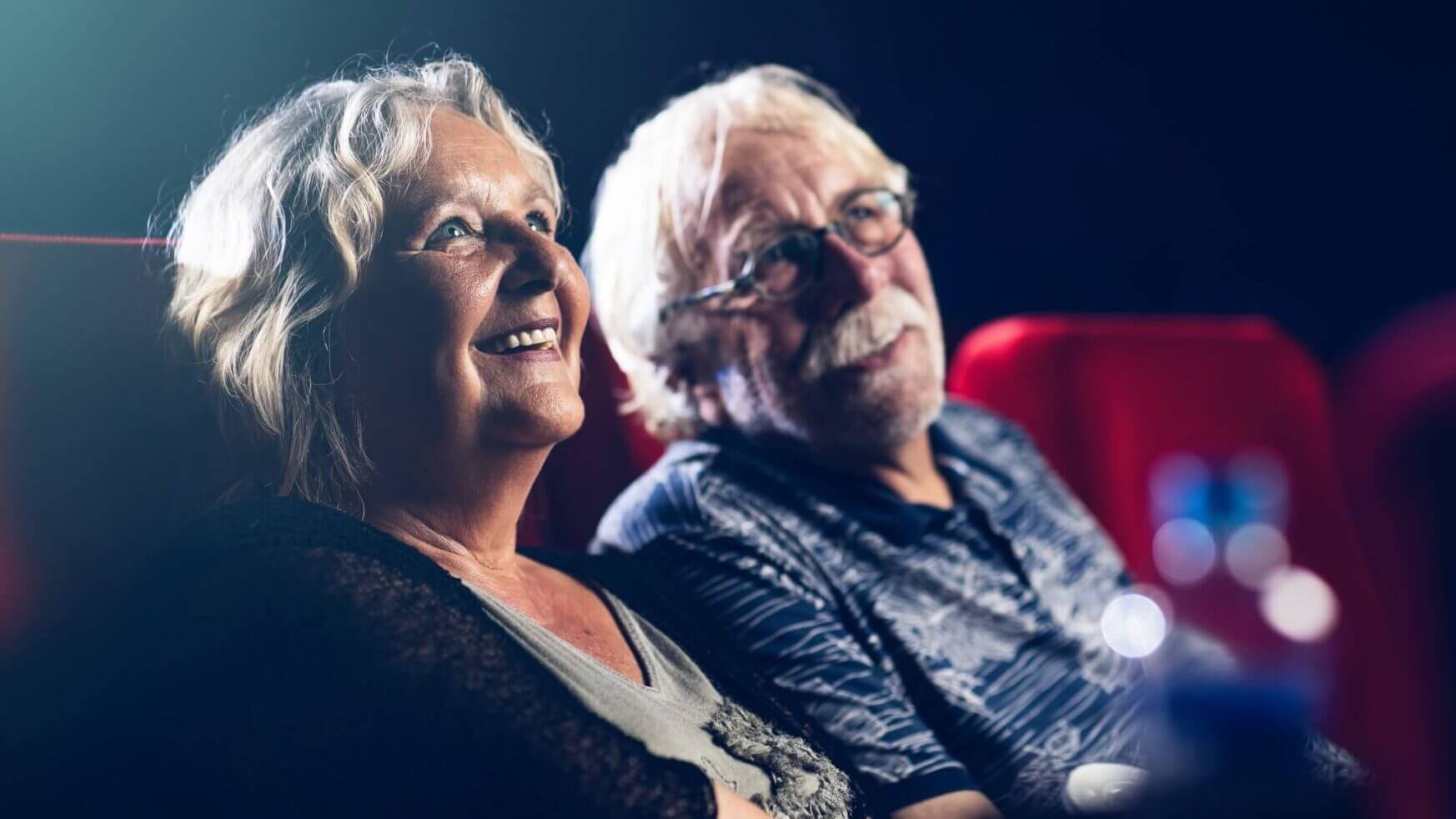 Een ouder echtpaar lacht terwijl ze genieten van een film in een theater met rode stoelen.