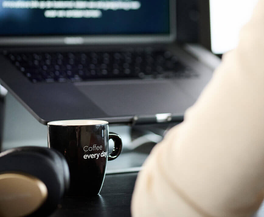 Een zwarte mok met de tekst "Koffie voor elke dag" staat op een bureau, naast een laptop en een koptelefoon.