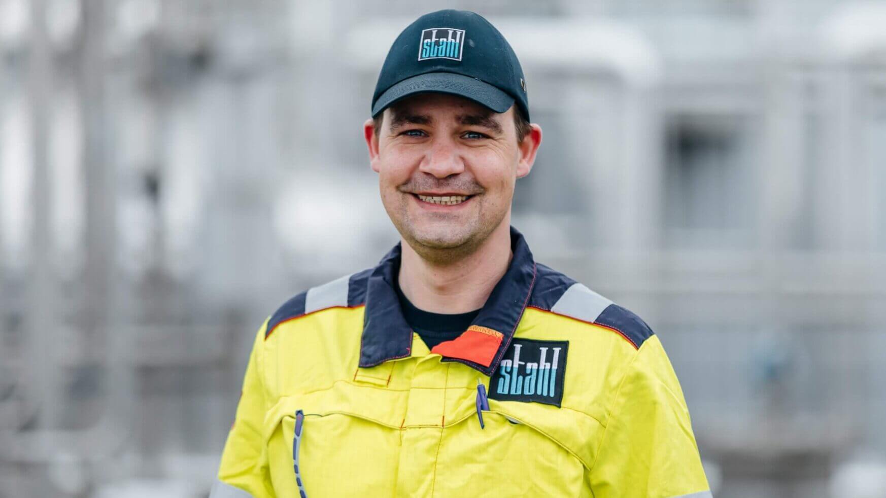 Een man lacht in een geel uniform en een zwarte Stahl-pet, tegen een industriële achtergrond.