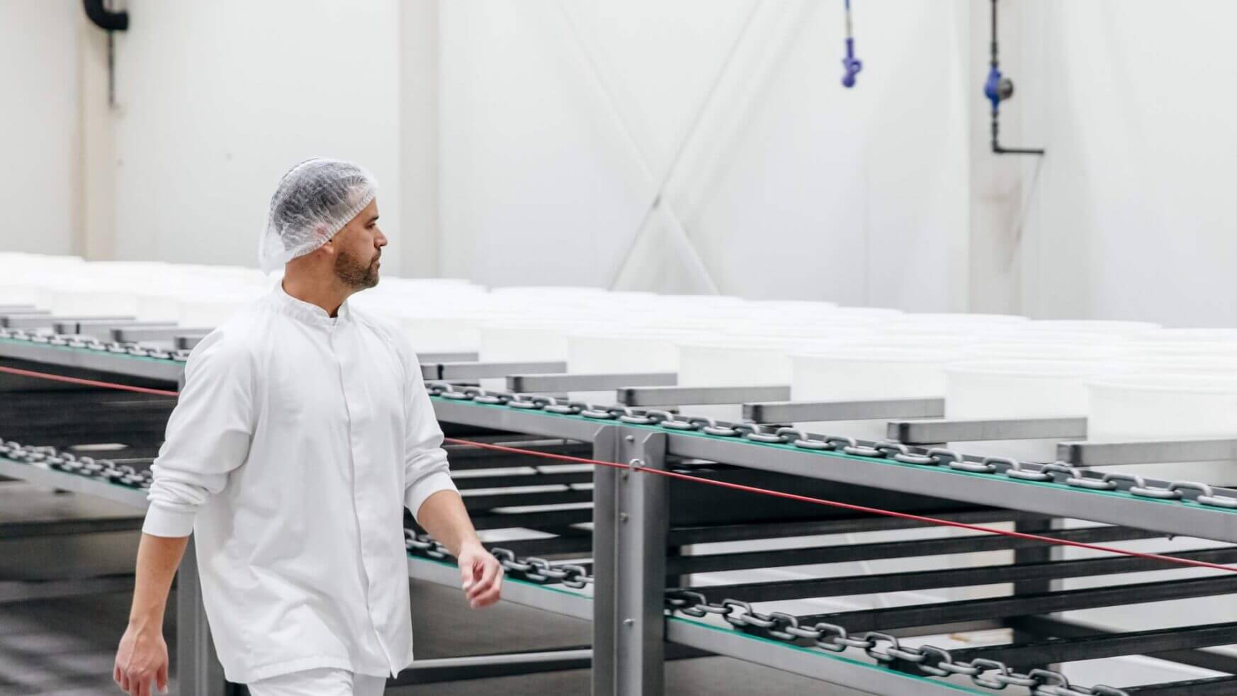 Een fabrieksarbeider in een wit uniform en met een haarnetje loopt langs transportbanden in een industriële omgeving.