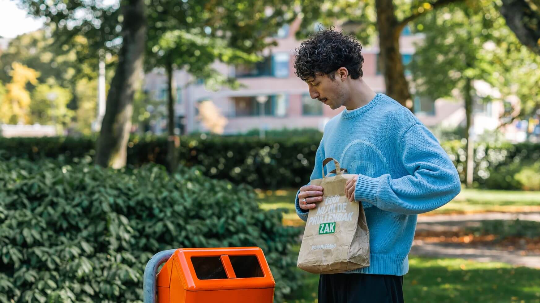 Man in blauwe trui gooit papieren zak in oranje recyclebak buiten-RSD aan de bak!.