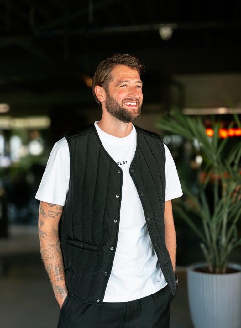 Een man met een baard, gekleed in een zwart vest en een wit T-shirt, lacht binnenshuis, omringd door planten.