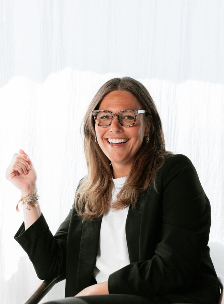 Een lachende vrouw met een bril en een zwart jasje zit tegen een lichte achtergrond.