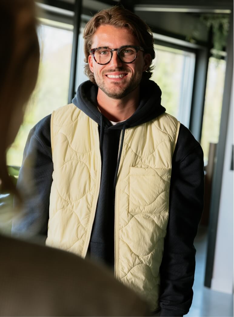 Lachende man met bril, die binnenshuis een crèmekleurig vest over een zwarte hoodie draagt.