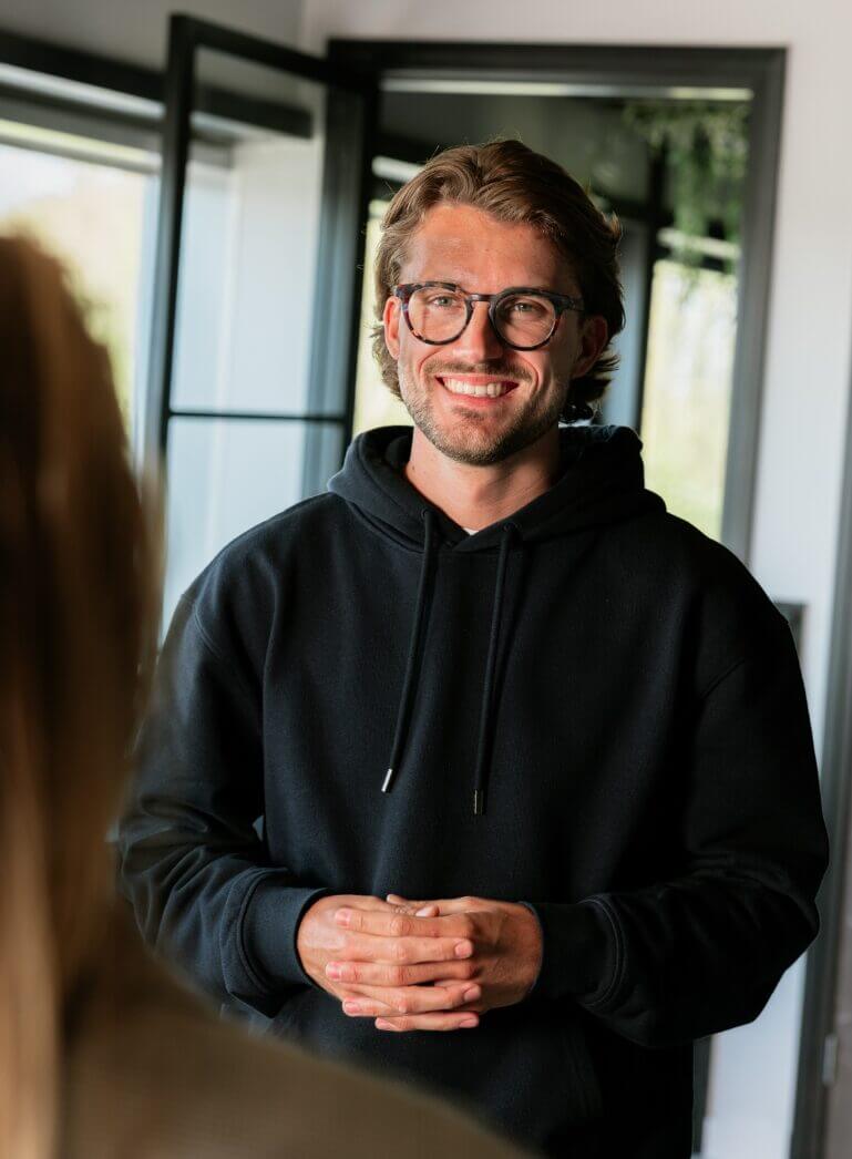 Een persoon met een bril en een zwarte hoodie lacht bij een open raam binnenshuis.