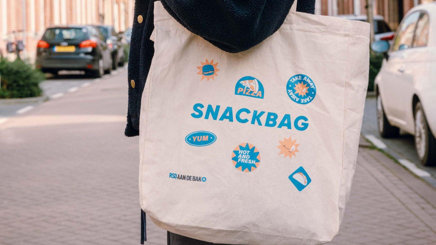 Persoon die door de stad loopt met een beige SNACKBAG tas bedekt met voedselstickers en een RSD aan de bak! badge.