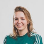 Blondine met schouderlang haar lacht in een groene Adidas-trui tegen een effen achtergrond.