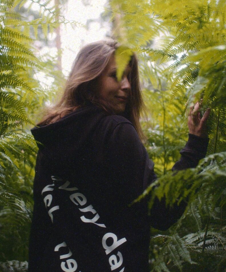 Vrouw in zwarte hoodie draait zich half om te midden van weelderige groene varens.