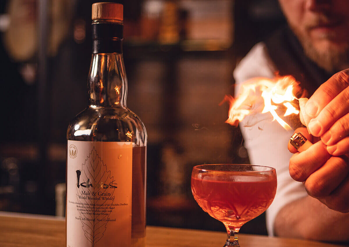 De barman steekt vlammen aan bij een cocktail, terwijl er een whiskyfles op de bar staat.