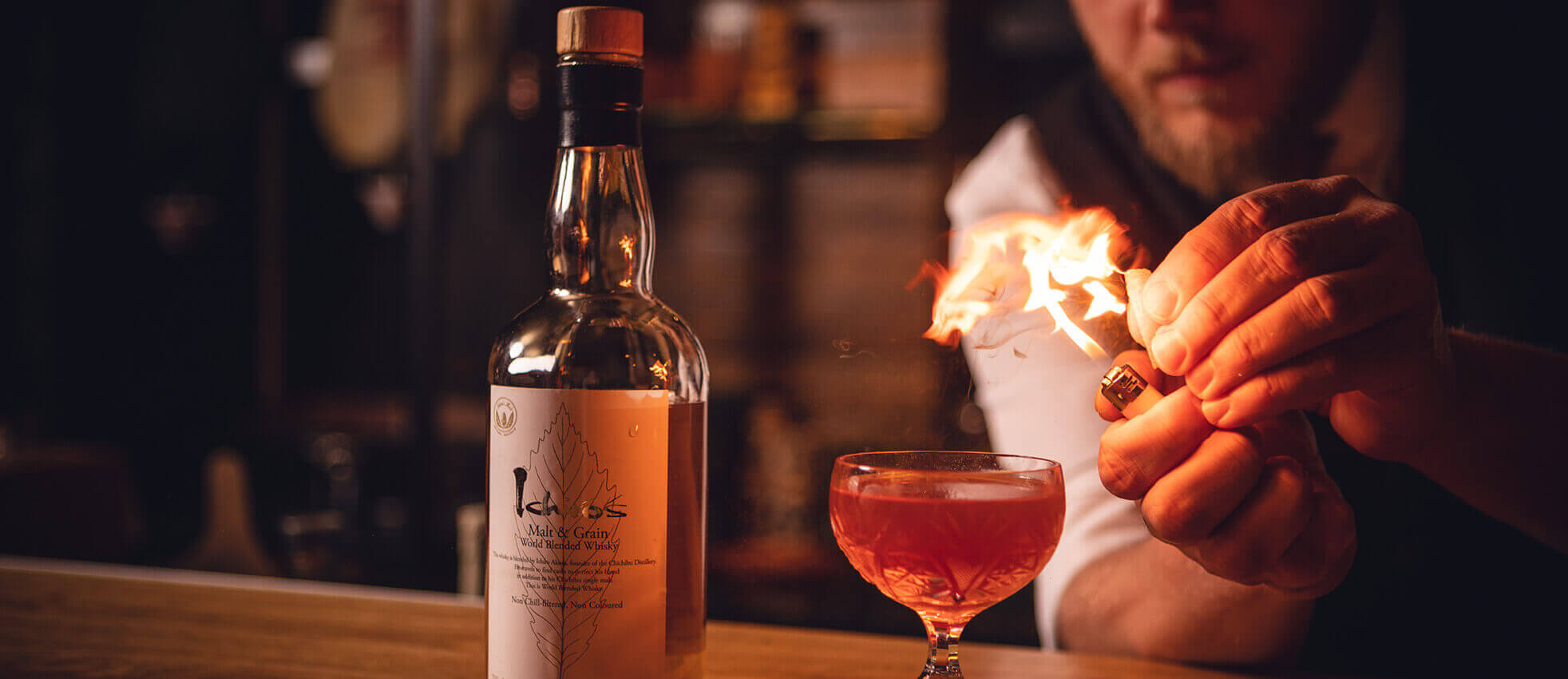 De barman steekt vlammen aan bij een cocktail, terwijl er een whiskyfles op de bar staat.