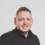 Kortharig persoon in een zwart shirt lacht tegen een eenvoudige achtergrond.