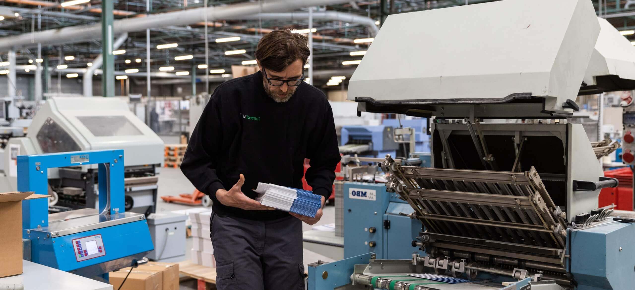 Een persoon beoordeelt afdrukken in een grote fabriek, omringd door allerlei drukmachines.