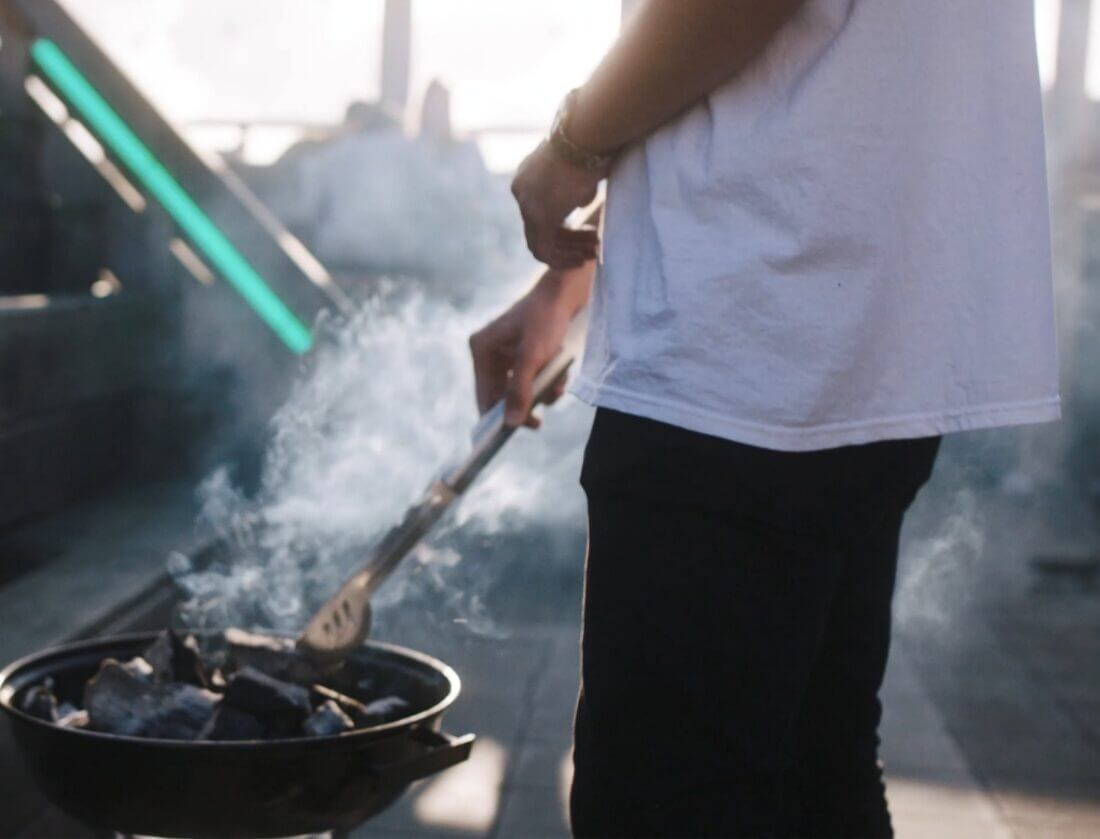 Grillen met een tang boven houtskool, gekleed in een wit shirt en een zwarte broek, terwijl de rook opstijgt.