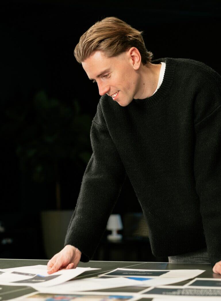 Een persoon in een zwarte trui bekijkt papieren op een tafel, tegen een donkere achtergrond.