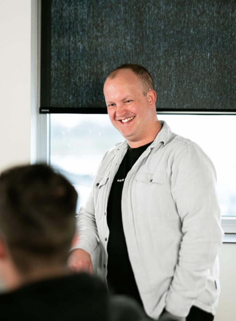 Een glimlachend persoon staat bij een raam met een zwart gordijn, te kletsen met iemand die vlakbij zit.