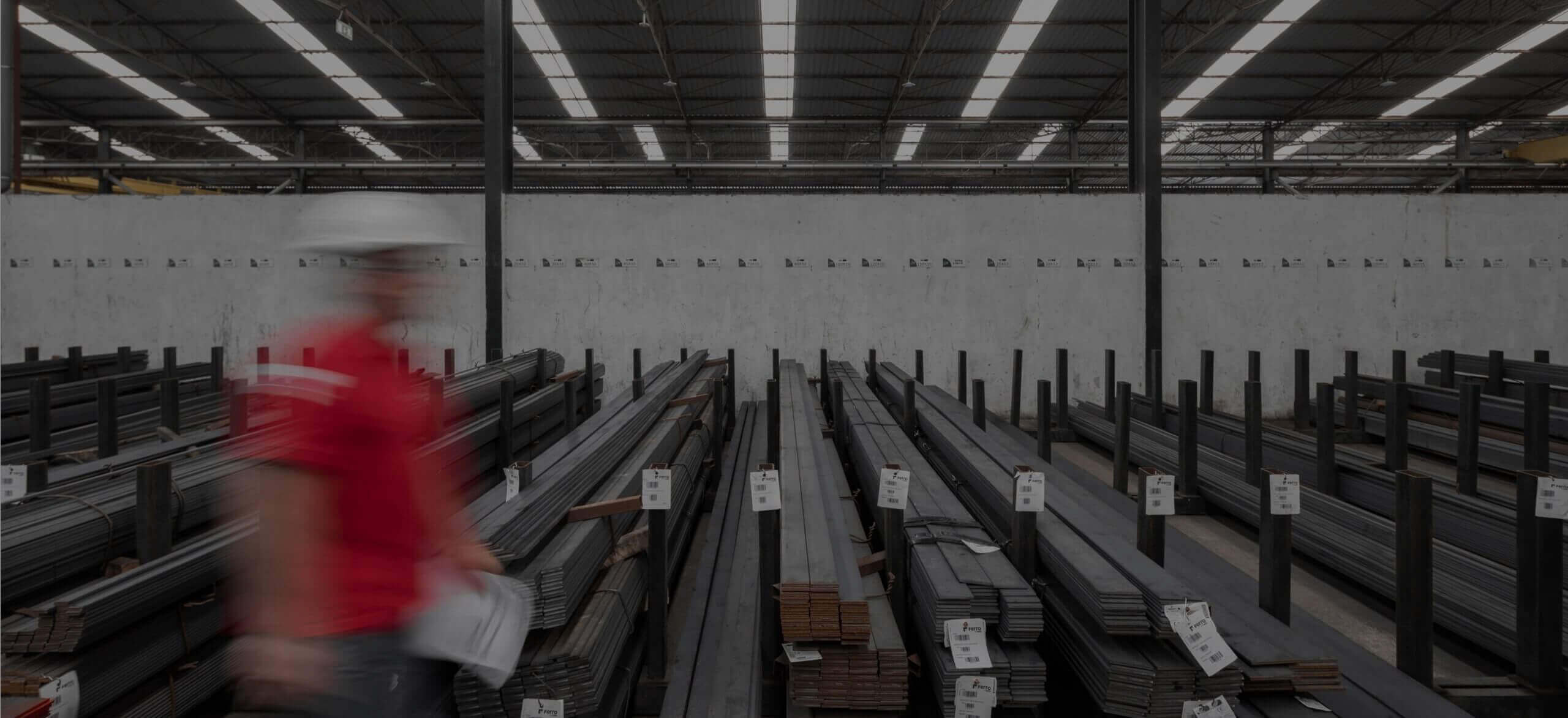 Pakhuis met opgestapelde metalen balken; links loopt een man in een rood jasje en een witte helm.