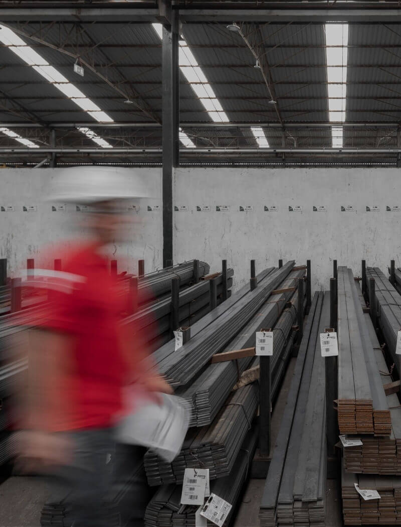 Een arbeider in een rood shirt en een helm loopt langs stapels metalen balken in een magazijn.
