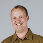 Persoon in een kaki shirt die lacht tegen een effen achtergrond.