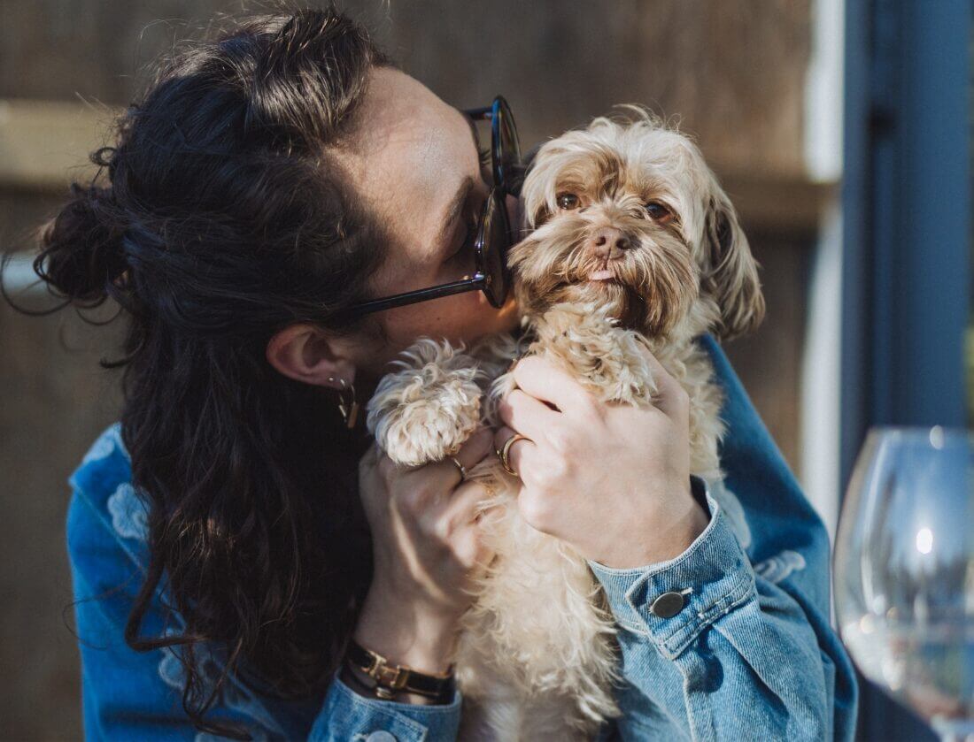 Vrouw met zonnebril, pluizig hondje in de hand, wijnglas in de buurt.