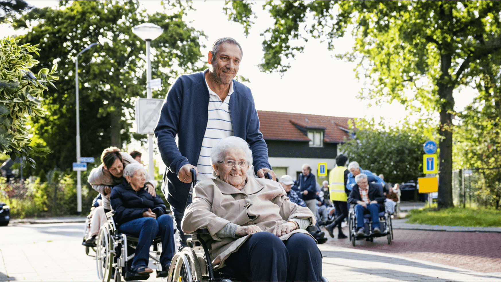 Een verzorger van Groenhuysen rijdt vrolijk een lachende oudere vrouw door een zonnige, levendige straat met veel groen.