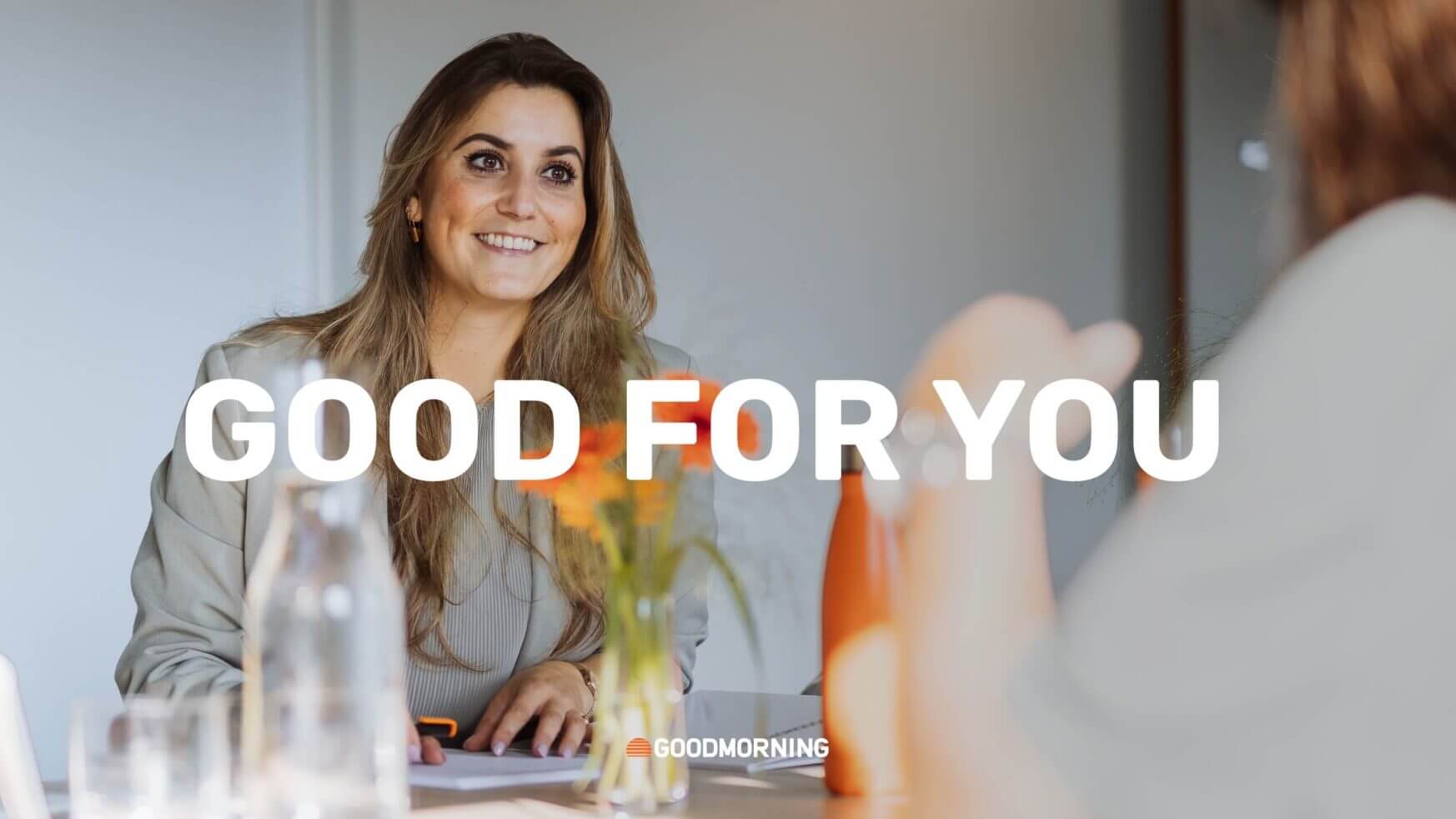 Een vrouw lacht aan haar bureau, bloemen en een fles water naast haar. Tekstoverlay: "GOED VOOR JOU.