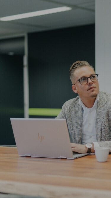 Een man met een bril en een blazer zit achter zijn bureau, zijn laptop open en kijkt peinzend omhoog.