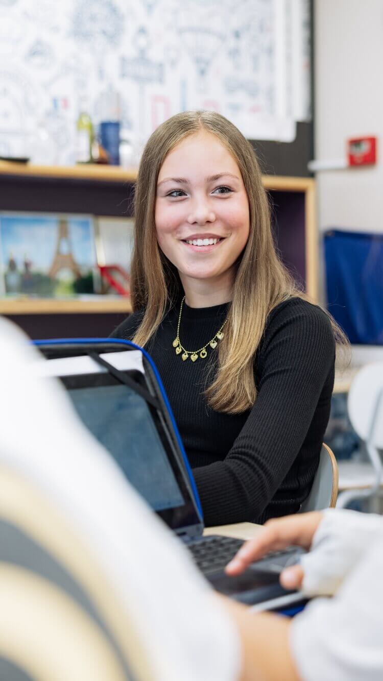 Een persoon in een zwart topje en een gouden ketting zit lachend met een open laptop in een klaslokaal.