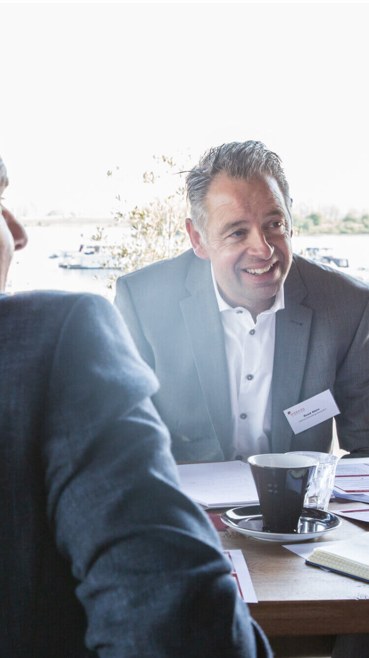 Een man in pak glimlacht aan een tafel met papieren en koffie in een lichte ruimte.