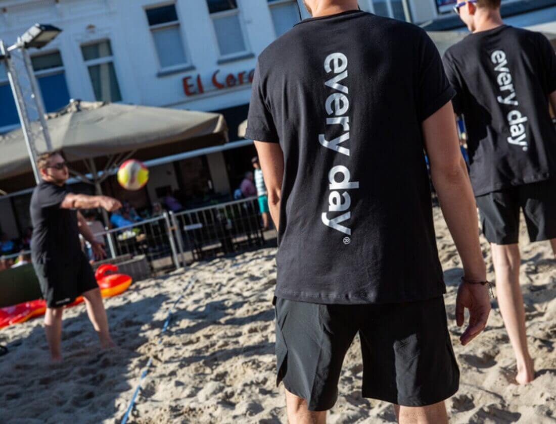 Spelers in zwarte T-shirts spelen volleybal op een zanderig terrein bij een gebouw.