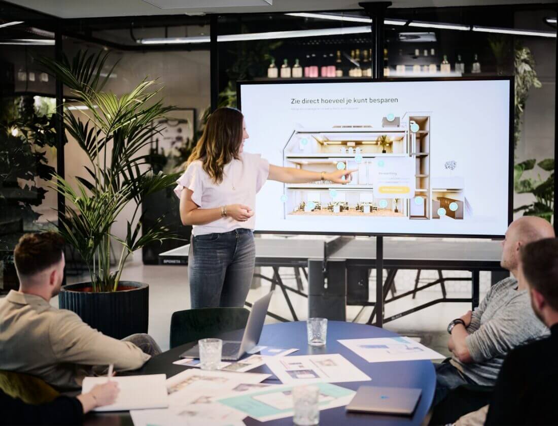 Een vrouw presenteert haar project op een scherm aan drie collega's in een modern kantoor.