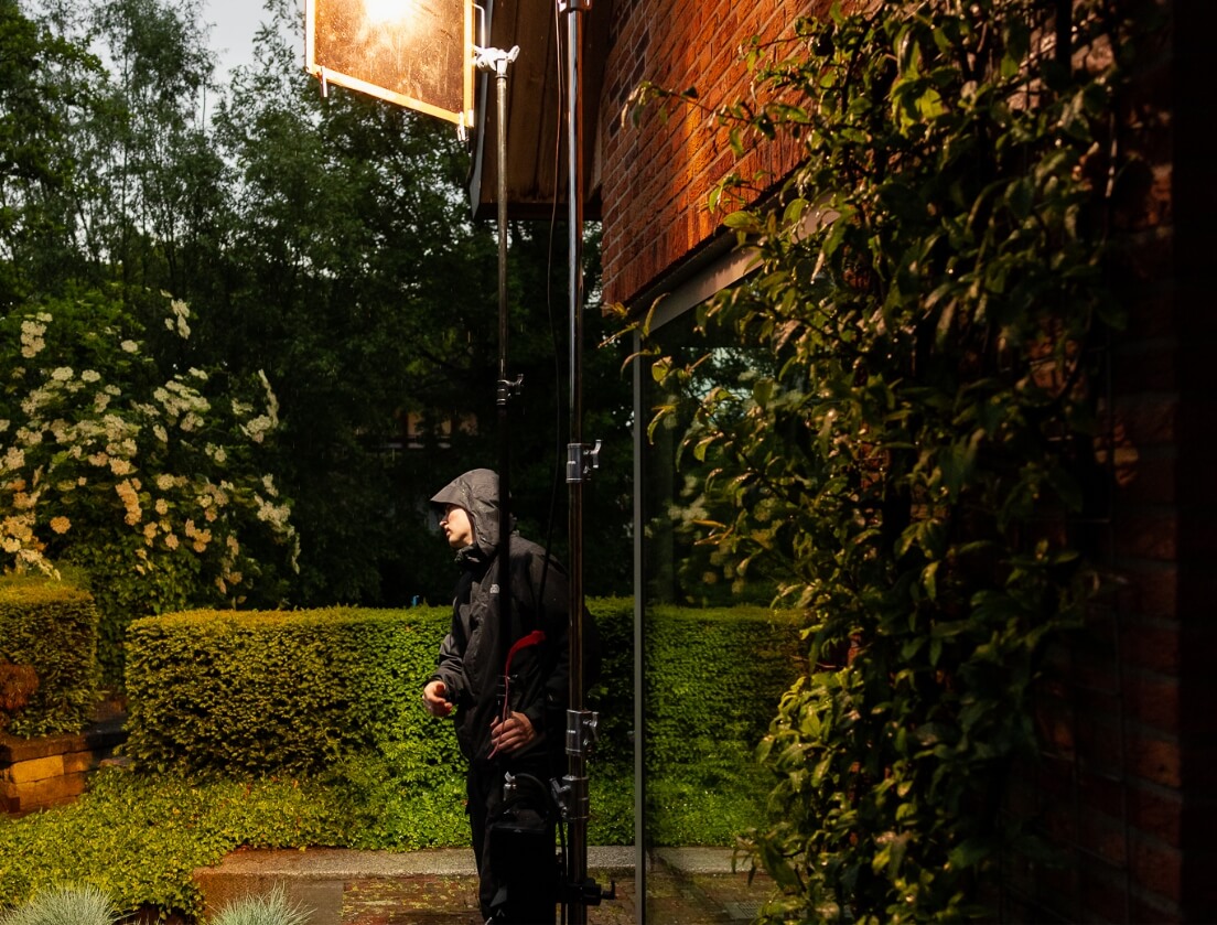 Figuur in regenjas naast filmapparatuur in een weelderige groene omgeving met een bakstenen muur.