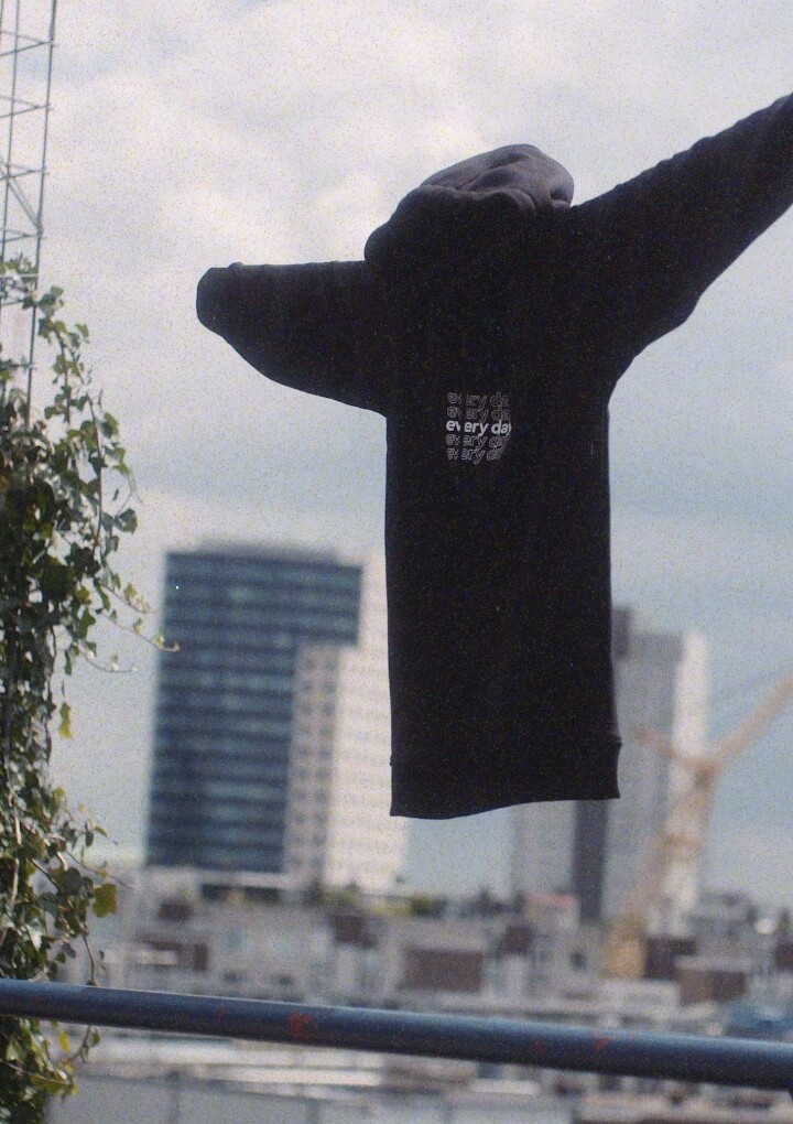 Zwarte hoodie met uitgestrekte armen; stadsgezicht, kraanvogel en klimop op de achtergrond.