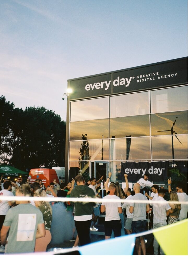 Zonsondergangfeest bij een modern gebouw, kleurrijke slingers, levendige menigte.