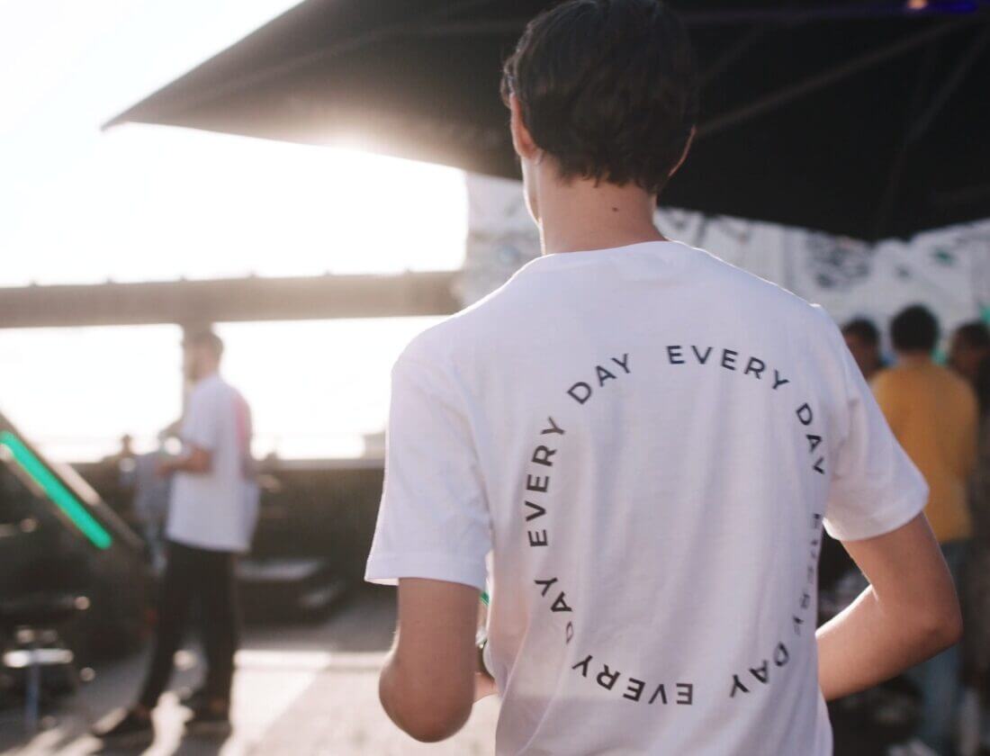 Een persoon in een wit "Every Day"-T-shirt loopt op een zonnige dag buiten, anderen op de achtergrond.