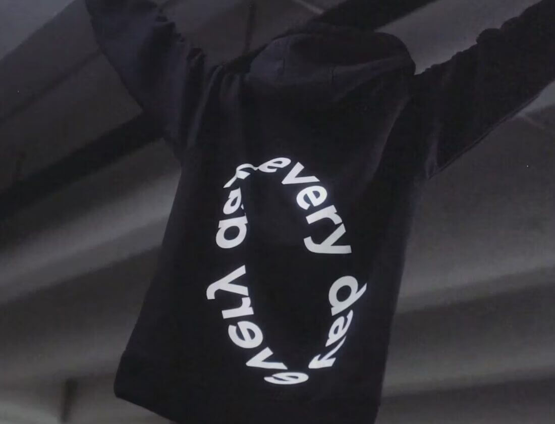 Een persoon in een zwarte hoodie met op de achterkant de tekst 'every day' in cirkelvorm, houdt zijn armen omhoog.