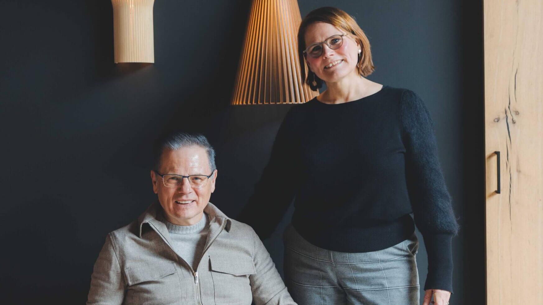 Lachend duo, één zittend en één staand, in een chique kamer verlicht door moderne TunnelmaDesign-lampen op donkere muren.