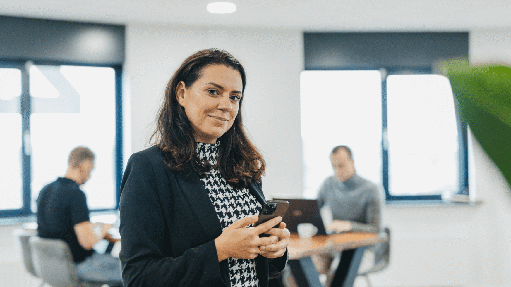 Een vrouw in een blazer glimlacht terwijl ze een telefoon vasthoudt, met twee collega's achter haar in een modern kantoor.