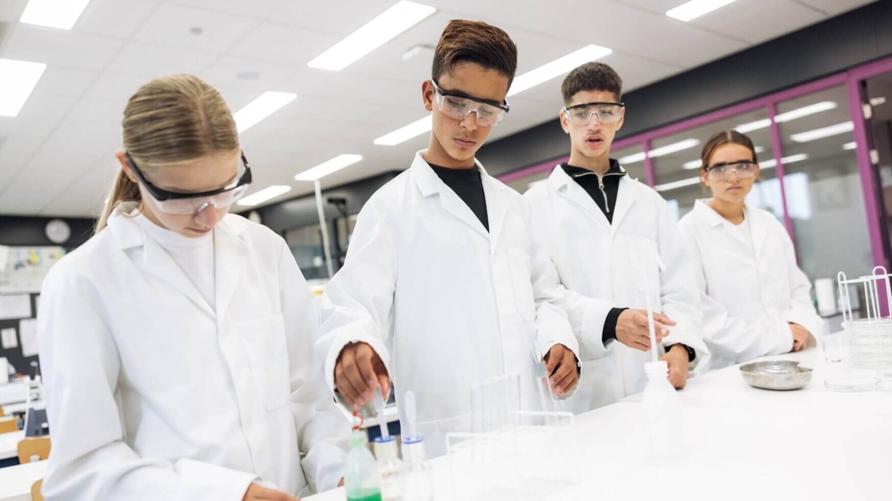 Vier studenten in laboratoriumjassen en met bril op experimenteren met reageerbuisjes op een laboratoriumtafel.