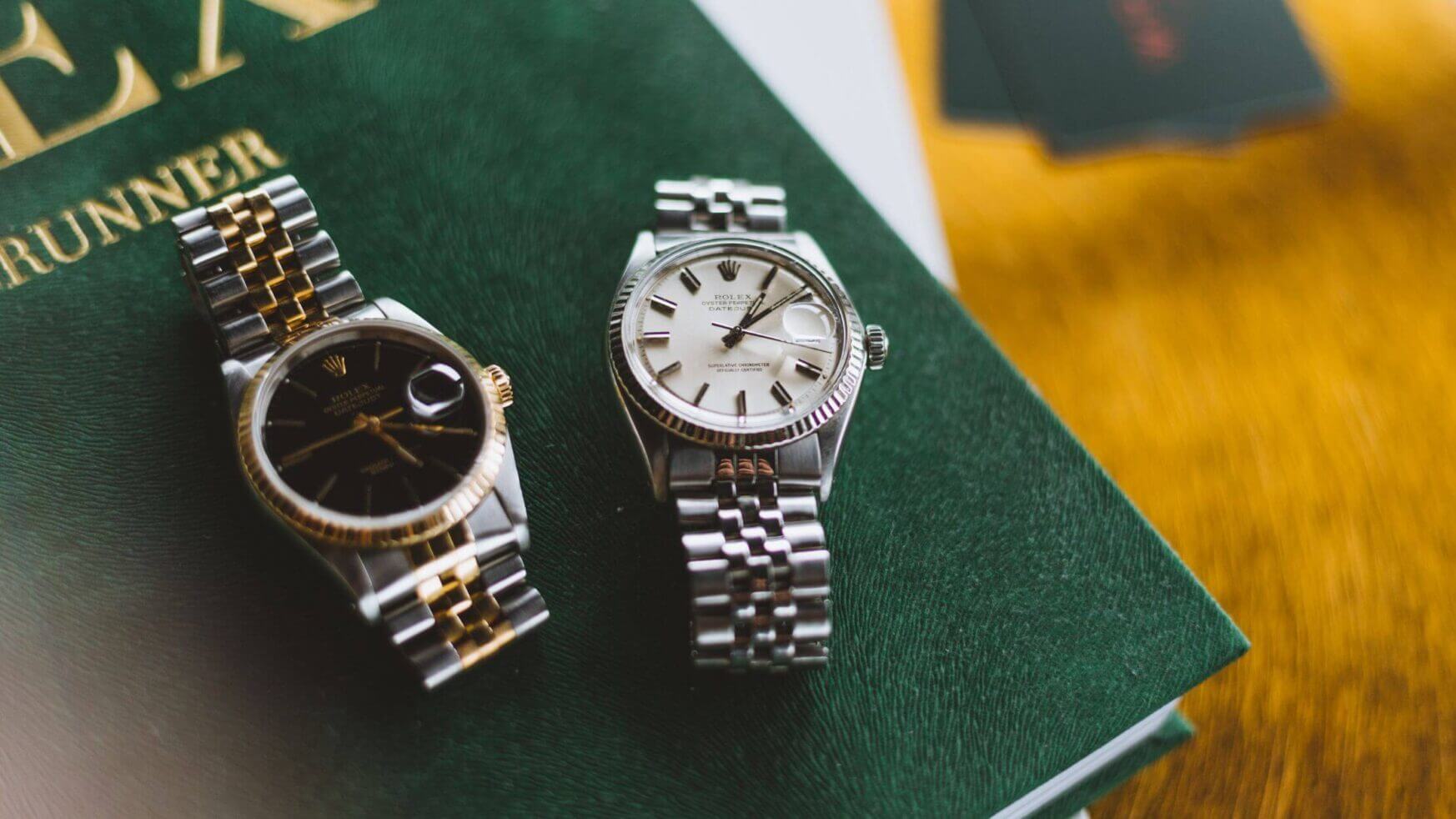 Op een groen boek liggen twee luxe metalen horloges: één met een zwarte wijzerplaat, de ander met een witte.