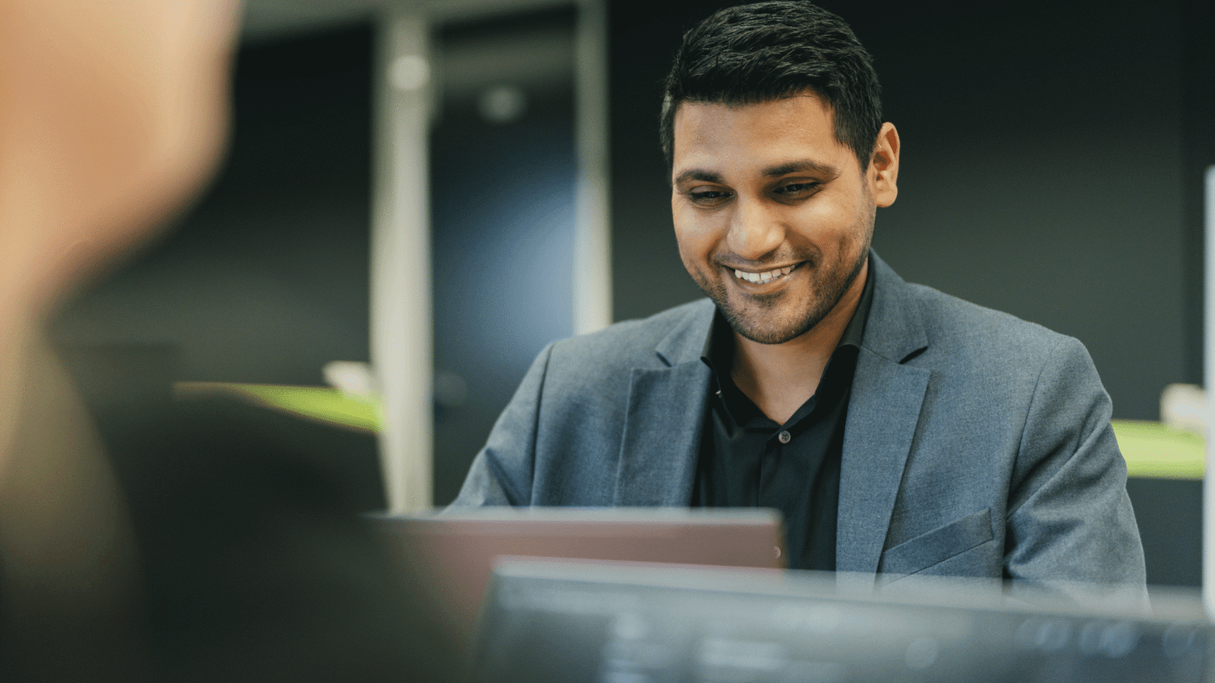 Man in pak, glimlachend zittend op kantoor en werkend op laptop.