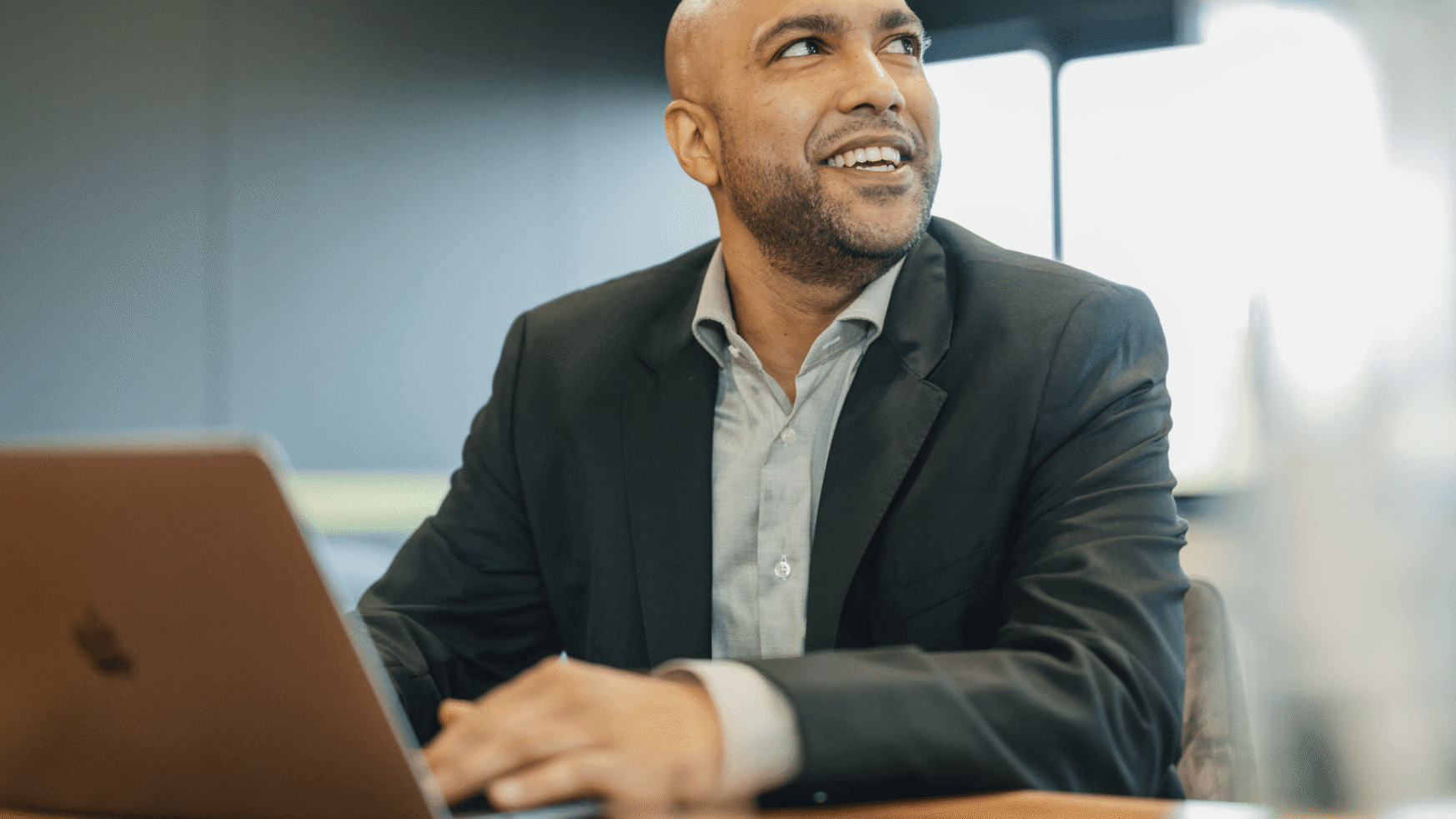 Een glimlachende man in pak werkt op zijn laptop aan een bureau.