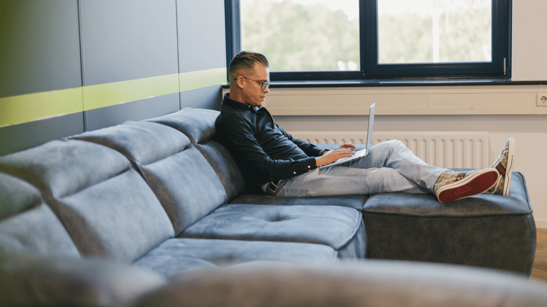 Ze zitten op een grijze bank bij het raam en werken op een laptop.