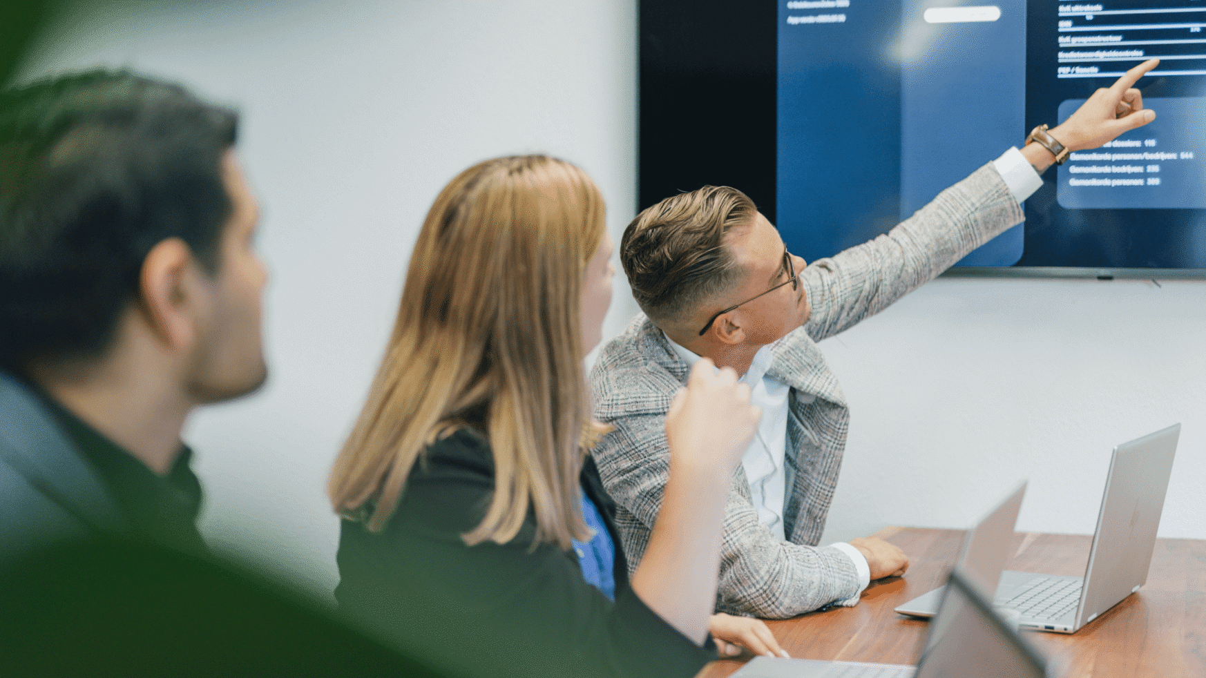 Drie mensen in vergadering, hun laptops open; één persoon wijst naar gegevens op het scherm.
