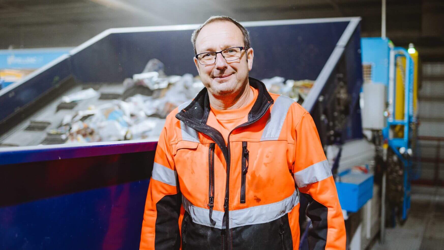 Een man in een oranje veiligheidsvest staat bij een recyclingmachine met daarin allerlei materialen.