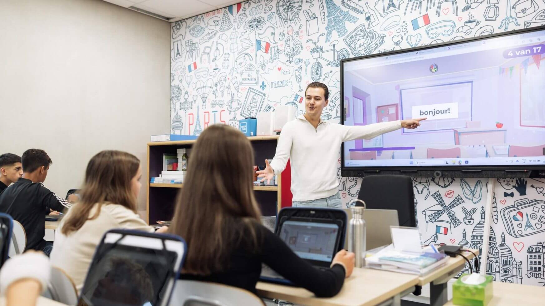 Een leraar wijst naar een "bonjour" op het scherm, terwijl leerlingen op laptops werken.